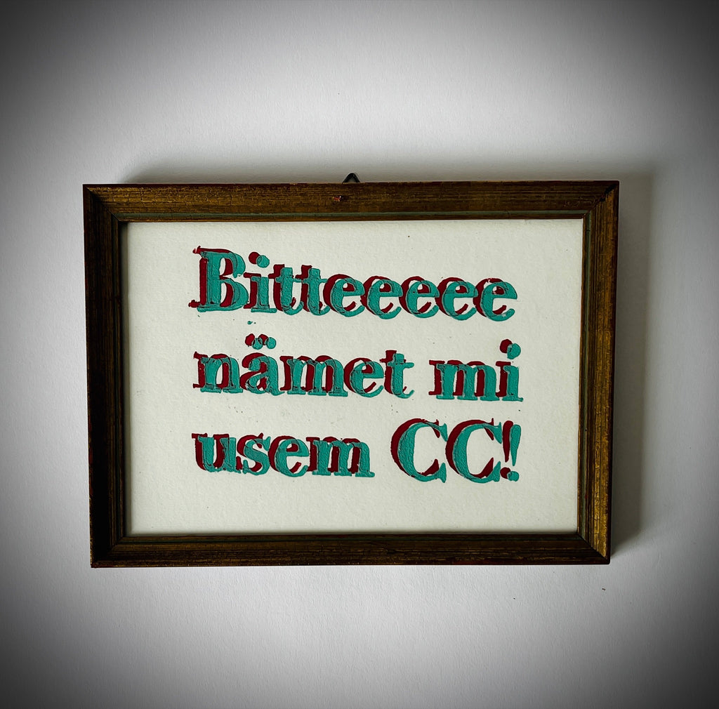 Gerahmter Linoldruck mit dem Schriftzug «Bitte nämet mi usem CC!» in Türkis und Dunkelrot, frontal an einer hellen Wand.
