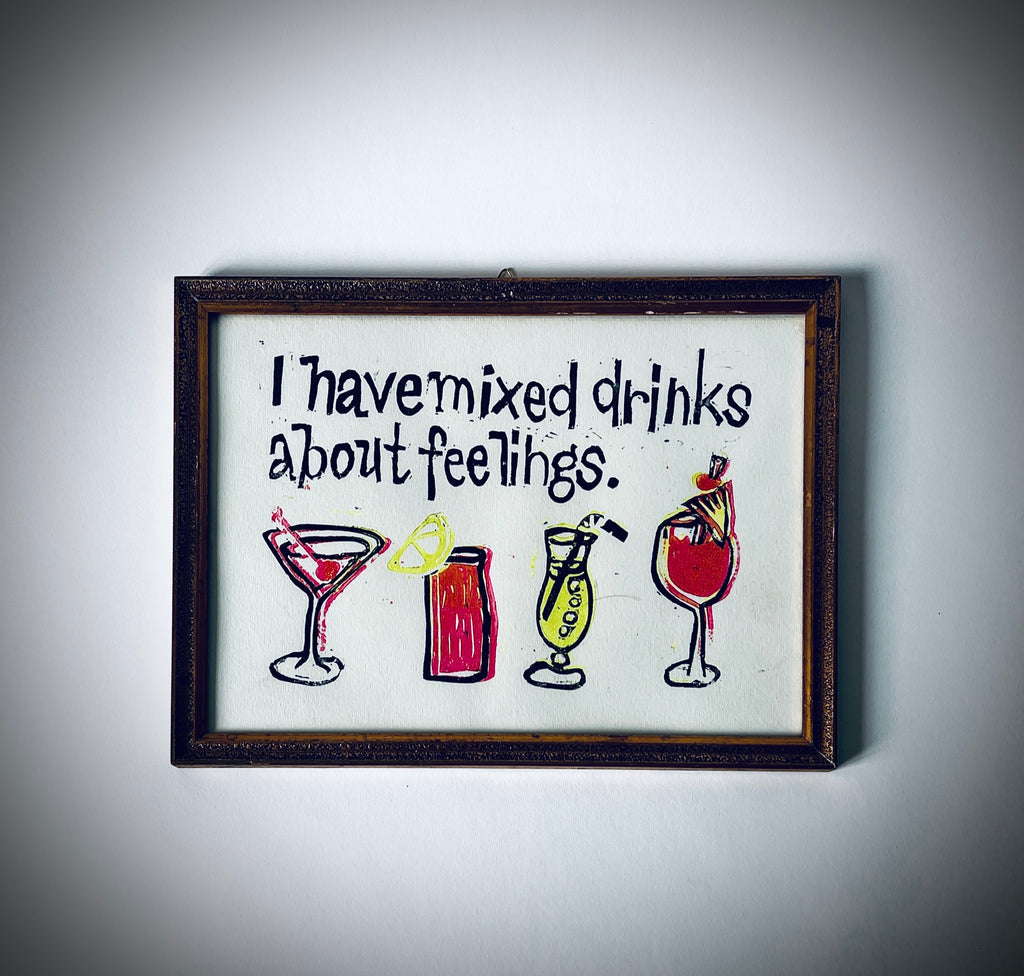 Gerahmter Linoldruck mit dem Satz «I have mixed drinks about feelings.» und vier Cocktailgläsern in dunklen Konturen, Rot und Gelb.