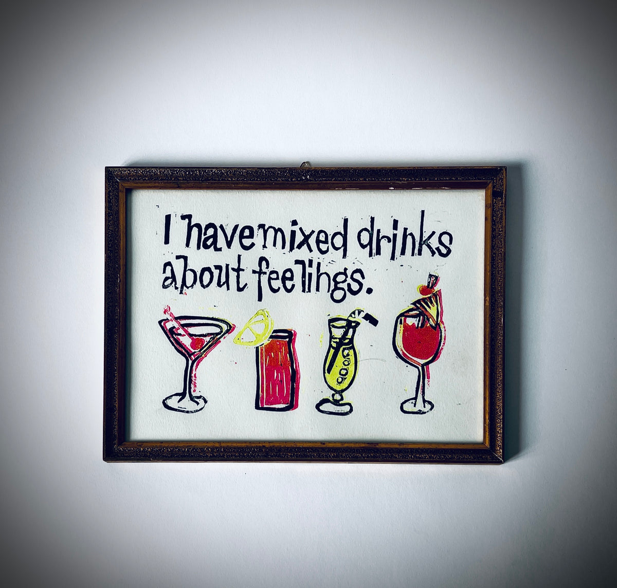 Gerahmter Linoldruck mit dem Satz «I have mixed drinks about feelings.» und vier Cocktailgläsern in dunklen Konturen, Rot und Gelb.