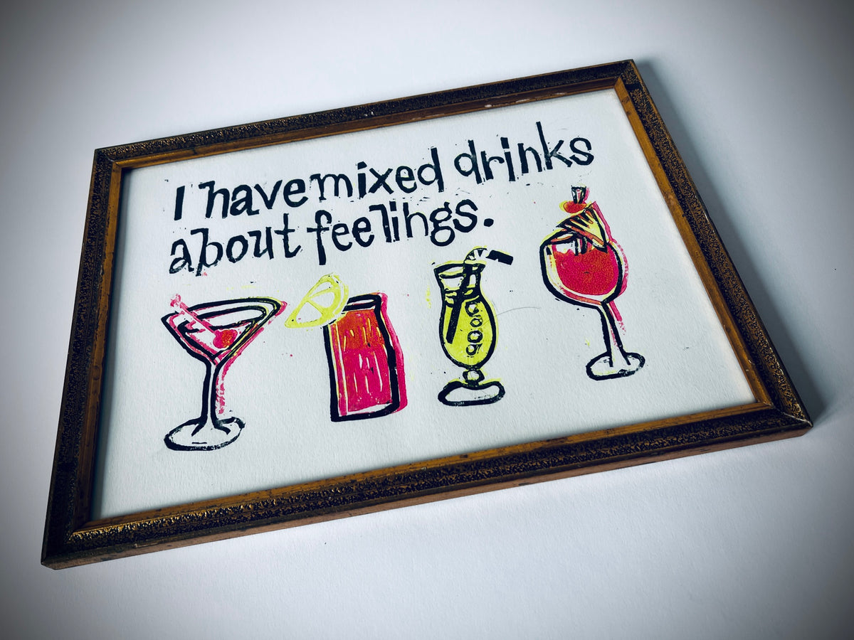 Schrägansicht des Linoldrucks «I have mixed drinks about feelings.» mit vier bunten Cocktailgläsern und dunkel-goldfarbenem Rahmen.