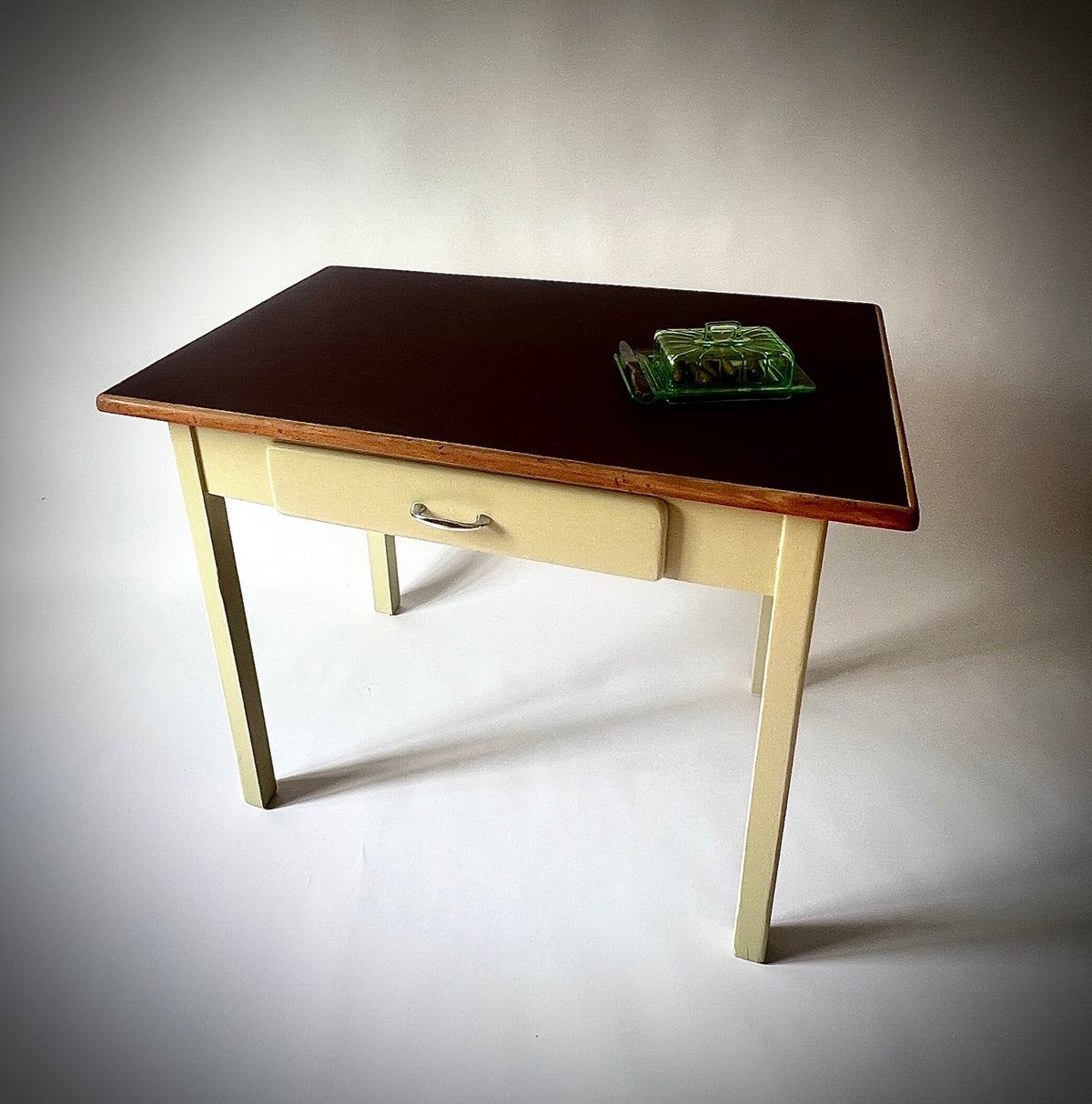 Tisch San Nazzaro mit dunkelroter Linoleumoberfläche, Holzumrandung, hellgelbem Untergestell und Schublade mit Metallgriff.
