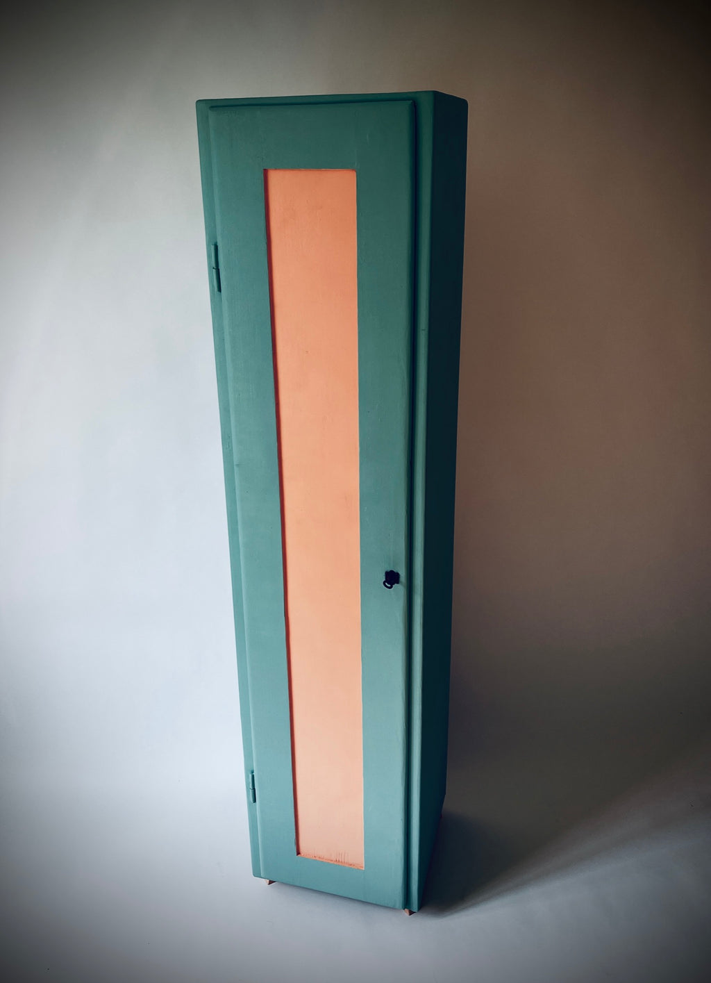 Schmaler Schrank S-chanf mit türkisgrünem Korpus, lachsfarbener Türfüllung und kleinen lachsfarbenen Füssen.
