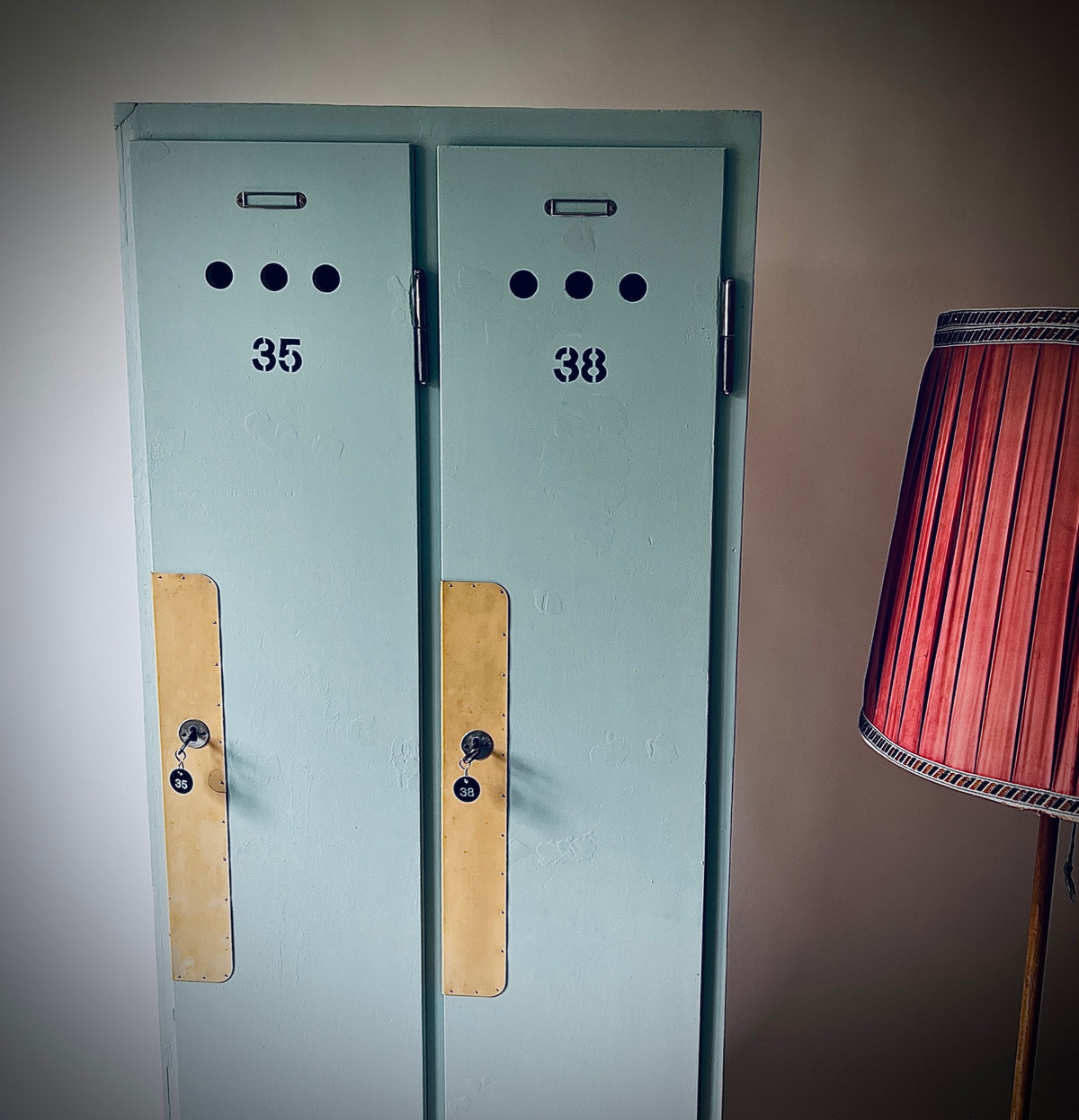 Blaugrüner Garderobenschrank mit zwei Türen, den Nummern 35 und 38, runden Lüftungslöchern und langen gelblichen Schutzplatten.