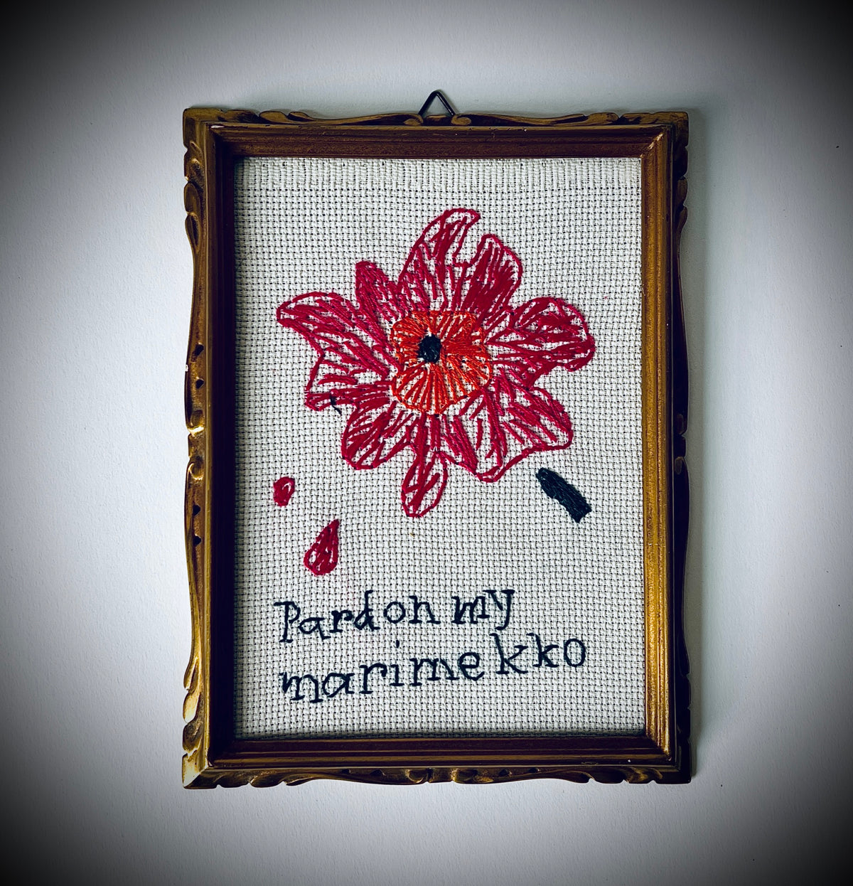 Gerahmte Stickerei mit pinkroter Blüte, orangefarbener Mitte und dunklem Schriftzug «pardon my marimekko» auf hellem Stoff.
