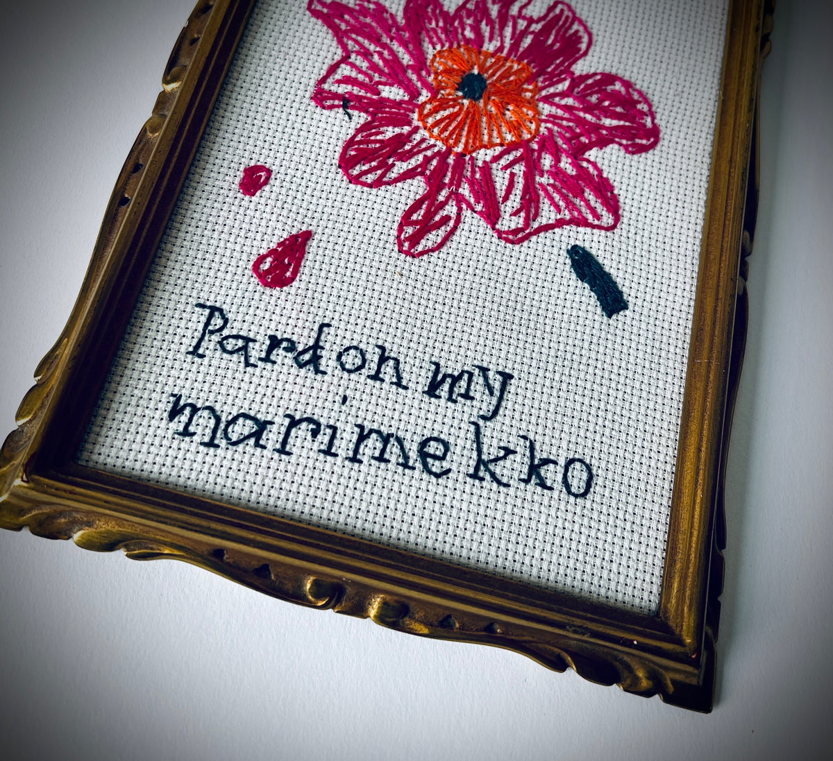 Nahaufnahme der gestickten Blüte und des Schriftzugs «pardon my marimekko» im goldfarbenen, verzierten Rahmen.
