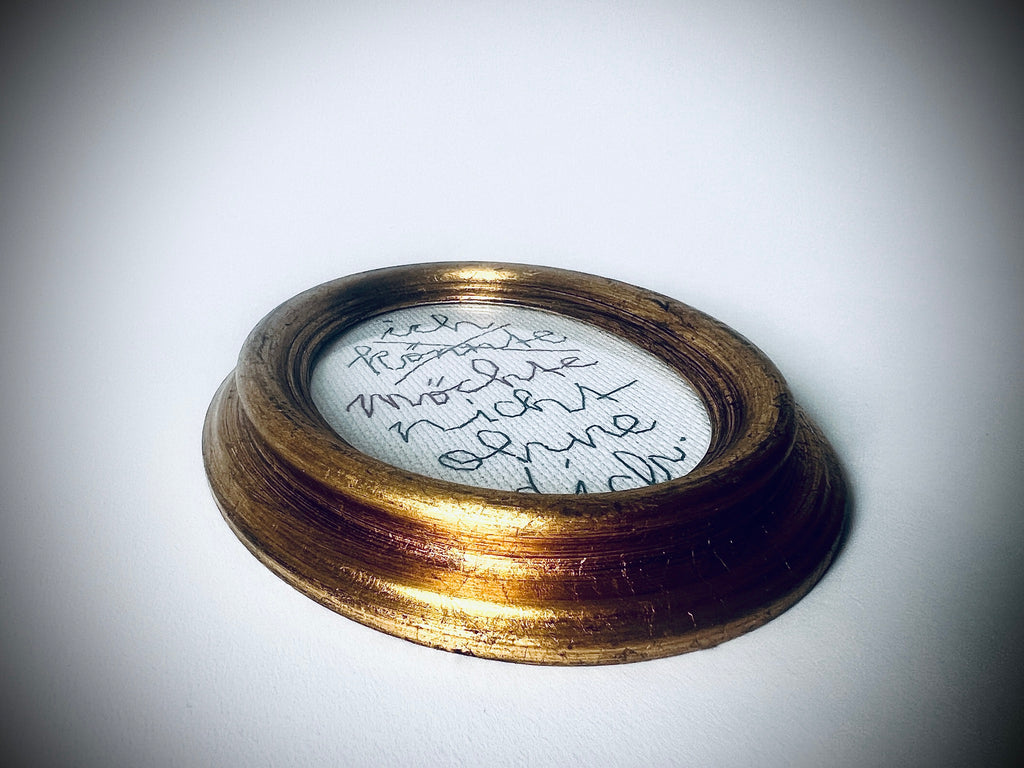 Flache Schrägansicht der Schriftstickerei «ich könnte/möchte nicht ohne dich» im goldfarbenen ovalen Rahmen.
