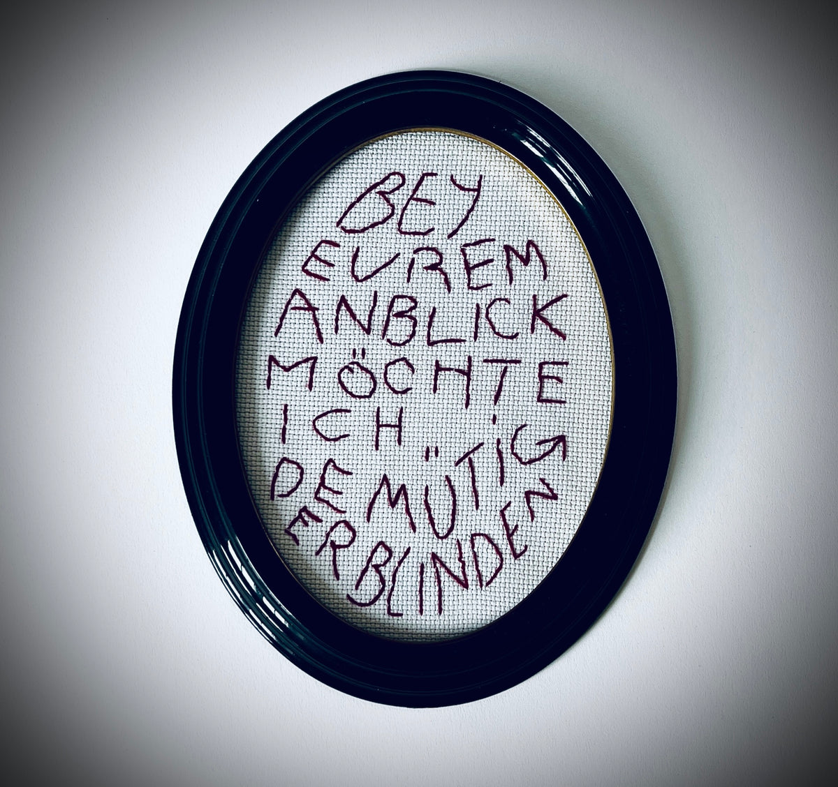Weinrote Schriftstickerei «Bey Eurem Anblick möchte ich demütig erblinden» in einem ovalen schwarzen Rahmen mit goldfarbener Innenkante.