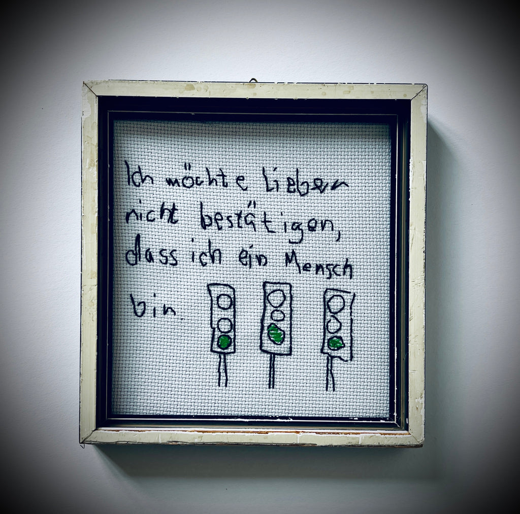Gerahmte Stickerei mit dem Satz «Ich möchte lieber nicht bestätigen, dass ich ein Mensch bin» und drei Ampeln mit grünem Licht.
