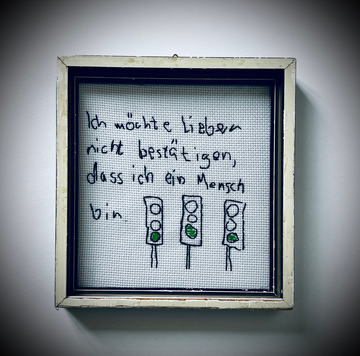 Gerahmte Stickerei mit dem Satz «Ich möchte lieber nicht bestätigen, dass ich ein Mensch bin» und drei Ampeln mit grünem Licht.
