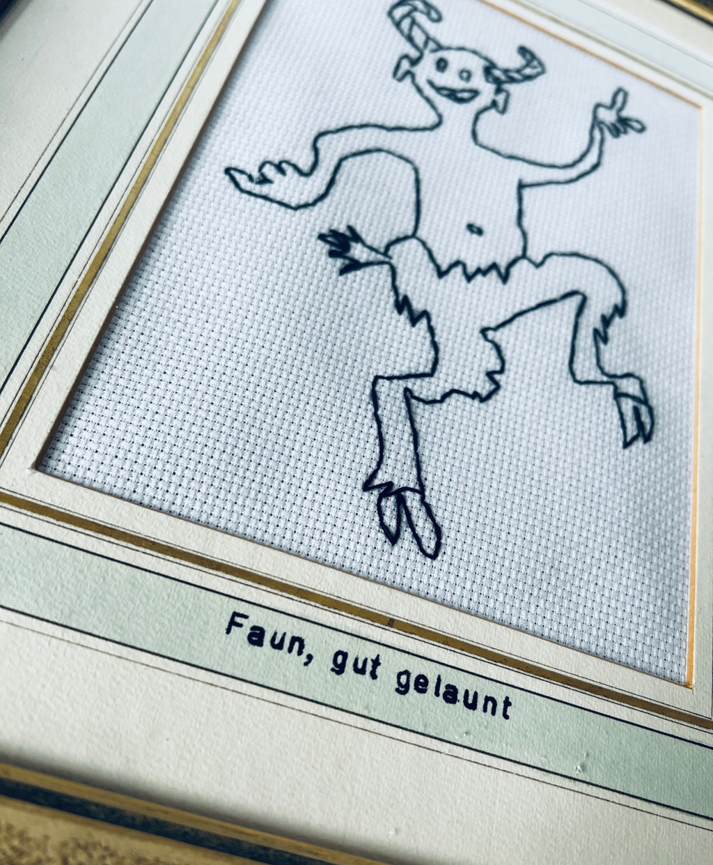 Nahaufnahme des gestickten Fauns und der Beschriftung «Faun, gut gelaunt» auf dem hellen Passepartout mit goldfarbenen Linien.