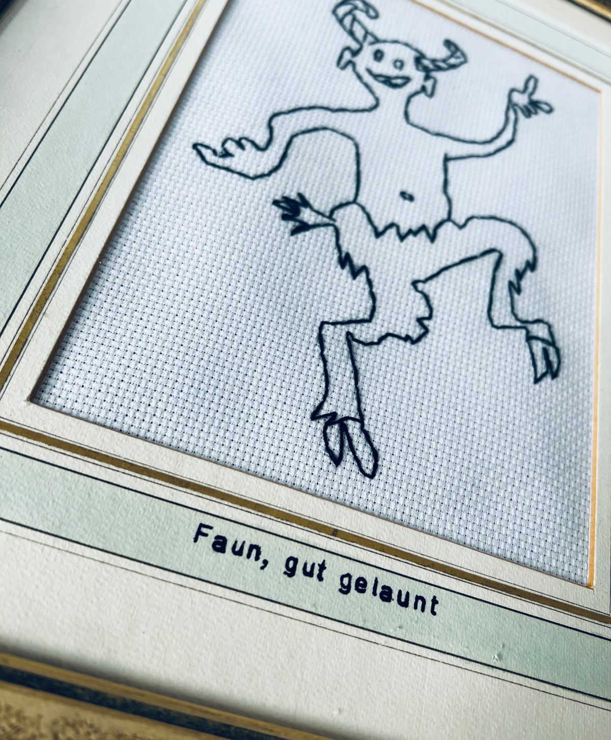 Nahaufnahme des gestickten Fauns und der Beschriftung «Faun, gut gelaunt» auf dem hellen Passepartout mit goldfarbenen Linien.